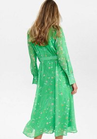 Lång grön klänning i genomskinligt tyg, med ett blommönster, långa ärmar och bälte i midjan. Kjolen slutar under knäet.