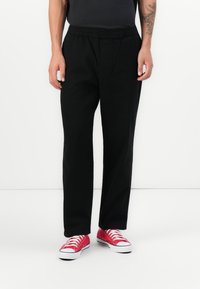 Person wearing black relaxed-fit pants and bright red low-top sneakers, standing against a plain white background.