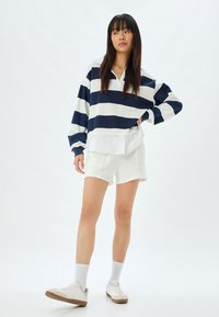 Pullover a righe blu navy e bianche con colletto, abbinato a pantaloni corti bianchi. La modella indossa sneaker bianche e calzini alla caviglia. Tessuto dal tocco morbido.