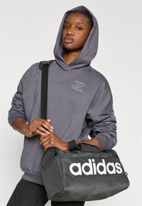 Pilkas džemperis su gobtuvu ir raišteliu, ant kurio yra atspausdintas logotipas, derinamas su juodu sportiniu krepšiu su didelėmis baltomis „adidas“ raidėmis.