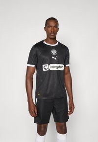 Maillot de sport noir avec des bordures blanches, arborant un logo circulaire et un texte de sponsor. Une paire de shorts noirs et des chaussettes blanches complètent le look.