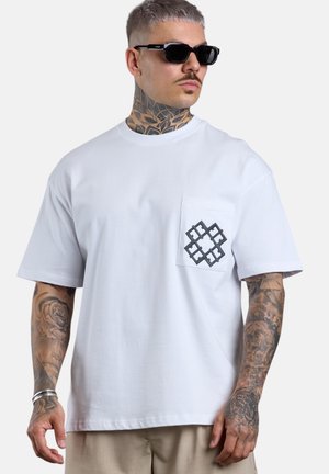 Man with tattoos wears black sunglasses and a white pocket t-shirt with a black geometric design on the pocket, beige pants, and silver bracelets.