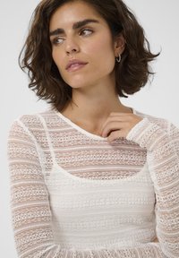 Weißes Spitzenoberteil mit langen Ärmeln, das mit komplizierten Mustern und Texturen versehen ist, mit einem Rundhalsausschnitt und enganliegenden Ärmeln.