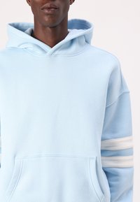 Světle modrá mikina s kapucí vyrobená z měkkého materiálu, s klokaní kapsou a třemi bílými pruhy na rukávech. Kapuce s šňůrkovým zavíráním.