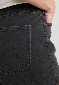Les jeans en denim noir présentent une poche arrière avec un motif de couture classique et un bouton en cuivre. Le tissu a une texture lisse.