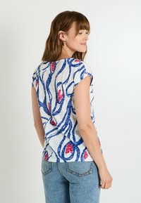 Top de manga corta en tejido ligero con un patrón floral en azul y rojo sobre un fondo blanco. Cuenta con un escote redondeado y un corte holgado.