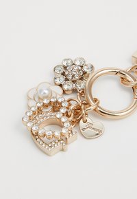 Portachiavi in tono dorato con un grande logo CC decorato con pietre di rhinestone trasparenti, un dettaglio a forma di fiore con centro in perla e un ciondolo a forma di cuore inciso con la parola "Guest."