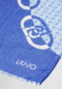 Sinine kangast sall, kus on geomeetriline muster ja logo. Materjal on pehme, lõpetatud frangeeritud Servadega. Erakordselt kontrastsed heledate ja tumedate siniste osade vahel.