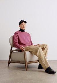 Maglione rosa chiaro, abbinato a un dolcevita nero, pantaloni beige e stivali neri. Seduto su una sedia in legno rotonda con siedi imbottito.
