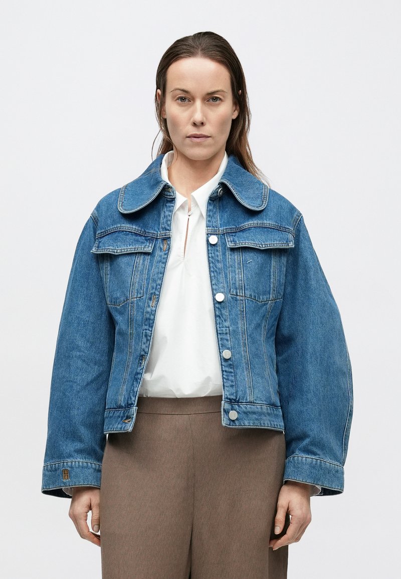 Blue denim jacket featuring a wide collar, front snap buttons, two chest pockets, and a relaxed fit. Smooth texture with visible seams.