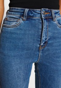 Vaqueros azules de denim con cierre frontal de cremallera y botón, usados con una camiseta negra visible en la cintura.