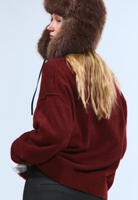 Pull bordeaux en tissu texturé avec ourlet côtelé, associé à un chapeau en fourrure marron avec rabats d'oreilles et cordon noir.