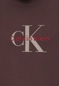 Bordó mikina s texturovanou látkou, vysokým límcem a velkým bílým logem "CK" s červenou výšivkou "CalvinKlein" pod ním.