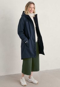 Marineblauwe waterdichte parka met capuchon, drukknoopsluiting, zijzakken, gedragen over een lichte trui en groene culottes, gecombineerd met witte sneakers.