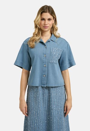 Woman wearing a light blue denim short-sleeve button-up shirt with sequined chest pocket and matching sequined skirt, facing forward.