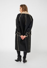 Svart overcoat med teksturert beige krage og beige sømmer. Har en løs passform, lange ermer, og når midt på leggen. Bæres med svarte bukser.