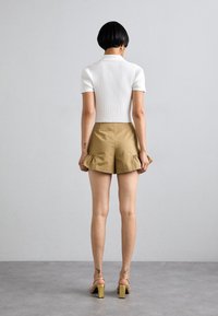 Vrouw met kort zwart haar in een witte ribgebreide top met korte mouwen, beige shorts met rimpelzoom, gouden oorbellen en gouden sandalen met hoge hakken.