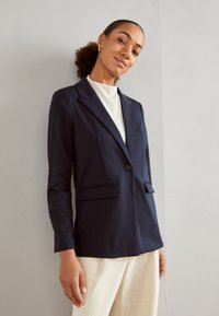 Blazer marine à revers, fermeture à un bouton, et deux poches avant, associé à un haut de couleur claire et un pantalon ivoire à motifs.