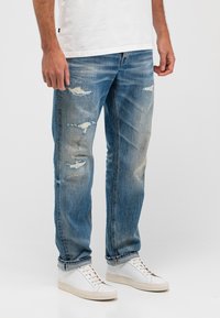 Mann trägt abgenutzte blaue Jeans, weiße Turnschuhe und ein weißes T-Shirt, steht vor einem schlichten hellen Hintergrund.
