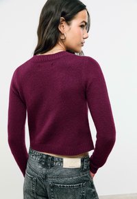 Femme tournée sur le côté, portant un pull cropped violet et un jean noir délavé taille haute avec une étiquette D66 sur la taille.