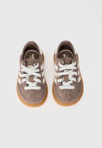 Par de ténis de camurça castanha com riscas brancas, atacadores e solas de borracha, vistos de cima sobre um fundo branco.