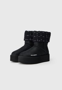 Botas negras de tobillo con un acabado brillante, un puño texturizado oscuro y una suela de plataforma gruesa. Presenta un detalle del logo en blanco en el lateral.