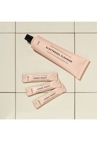 Electrocel-Reiniger in einer sanften pinkfarbenen Tube mit schwarzem Deckel, neben drei pinken Beuteln mit dem Aufdruck Ionic Shot, platziert auf einer hellen gefliesten Oberfläche.