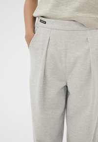 Világosszürke, redőzött nadrág, puha anyagból készült, fekete címkével ellátott derékkal és oldalsó zsebekkel a praktikumért.