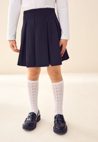 Jupe plissée noire, chaussettes blanches texturées montant jusqu'aux genoux, et mocassins noirs brillants. La tenue présente un design simple et classique.