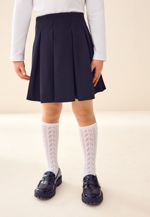 Gonna nera plissettata, calzini bianchi strutturati al ginocchio e mocassini neri lucidi. L'outfit presenta un design semplice e classico.