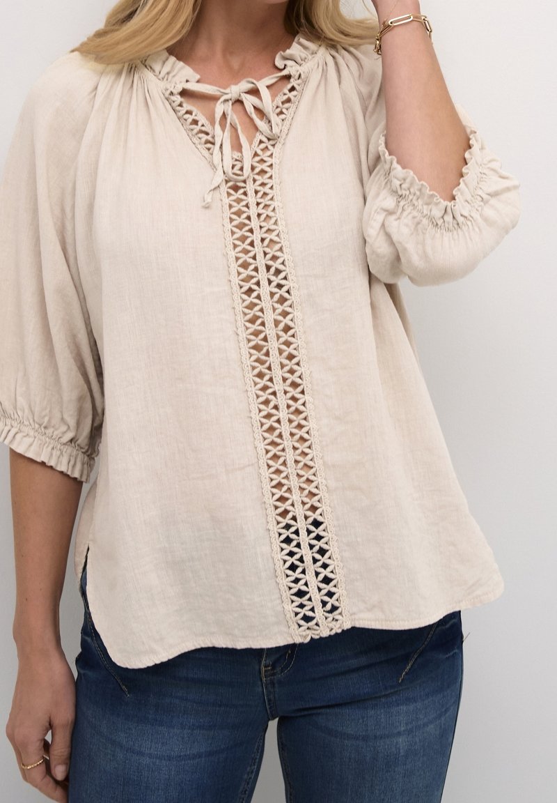 Blouse en lin beige avec un insert en dentelle sur le devant, encolure et manches froncées, et coupe décontractée, associée à un jean bleu.