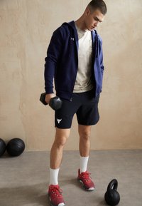 Marineblauwe hoodie met rits, beige t-shirt, zwarte shorts met wit logo, rode sneakers, houdend in de hand dumbbells, fitnessomgeving met gewichten in de buurt.