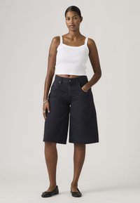 Γυναίκα όρθια με το ένα χέρι στην τσέπη, φορώντας λευκό crop top, μαύρα, ευρύχωρα σορτς μέχρι το γόνατο και μαύρα, επίπεδα παπούτσια, ουδέτερο φόντο.