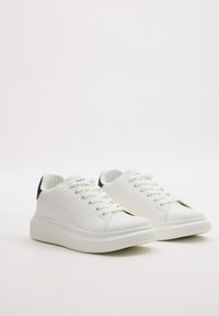 Zapatillas blancas con empeine de cuero liso, punta redonda, suela plana de goma blanca y acento azul oscuro en el talón. Presentan cordones y pequeñas perforaciones.