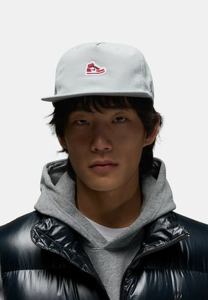 Cappello grigio con logo di sneaker rosso, indossato con una felpa grigia e un gilet imbottito nero lucido, con chiusura a zip e modello trapuntato.