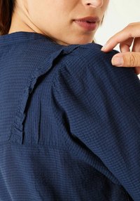 Marinblå blus med textur som har volangdetaljer vid axlarna och veckade ärmar. Tyget har ett subtilt rutmönster och känns mjukt vid beröring.