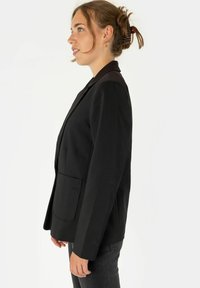 Črni blazer s strukturiranim dizajnom, enim sprednjim žepom, režastimi reverji in prilegajočim krojem. Gladek material in minimalni kovinski detajli.