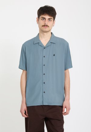 Giovane uomo con capelli corti e scuri e baffi, indossa una camicia blu a maniche corte con bottoni e pantaloni marrone scuro, in piedi davanti a uno sfondo semplice.