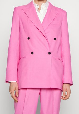 Osoba ubrana w jaskrawy różowy dwurzędowy blezer z czarnymi guzikami oraz dopasowane spodnie, na białej koszuli z zapięciem na guziki.