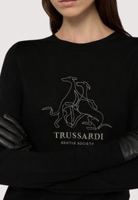 Svart tröja med ribbad struktur, med en vit konturdesign av hundar och "TRUSSARDI GENTLE SOCIETY"-logotypen nedanför. Svarta läderdetaljer.
