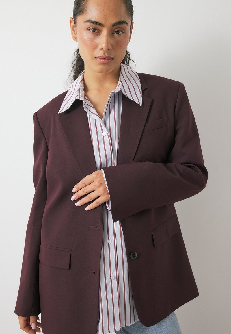 Donna con capelli scuri raccolti indossa un blazer oversize di colore viola scuro sopra una camicia bianca a righe rosse, in piedi davanti a un muro bianco semplice.