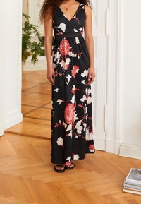 Černé maxi šaty s červeným a bílým květinovým vzorem, v-výstřih, lehká tkanina a přiléhavý pas. Modelka má na sobě černé sandály.