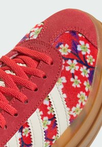 Rote Wildleder-Sneaker mit weißen Streifen, ausgestattet mit einem floralen Design in Rot, Pink und Grün an der Seite und strukturierten Schnürsenkeln. Durchsichtige Gummisohle.