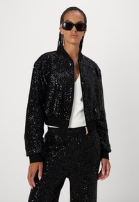 Black sequined bomber jacket with a ribbed collar and cuffs, zip closure, and a cropped design. Worn with matching sequined pants.
