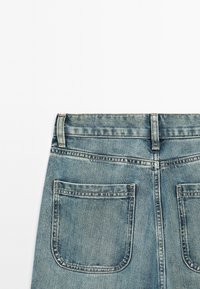 Zadní pohled na modré džíny z denimu, ukazující poutka na opasek, švy a dvě čtvercové zadní kapsy na bílém pozadí.