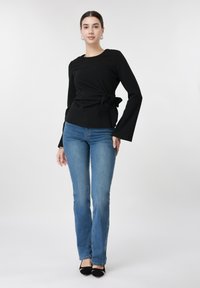 Γυναίκα που φοράει μαύρη μπλούζα με μακριά μανίκια και δέσιμο στο πλάι, μπλε τζιν, μαύρα μυτερά παπούτσια, στέκεται μπροστά σε απλό λευκό φόντο.