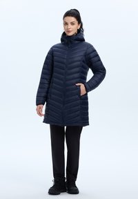 Laivastonsininen puffatakki, jossa on viisto muotoilu, vetoketjusuljenta, sivutaskut ja säädettävä huppu. Yhdistetty mustiin housuihin ja saappaisiin.