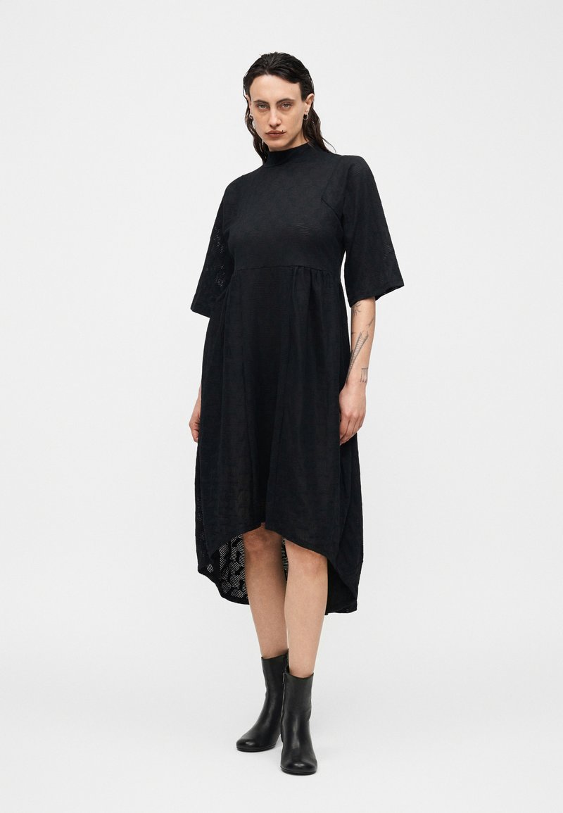 Frau trägt ein schwarzes Kleid mit hohem Kragen und Ellenbogenärmeln, asymmetrischem Saum und schwarzen Stiefeletten, stehend vor weißem Hintergrund.