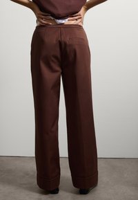 Pantalons ample en marron avec une texture lisse, dotés d'une poche arrière et de poignets repliés. La taille est conçue pour le confort.