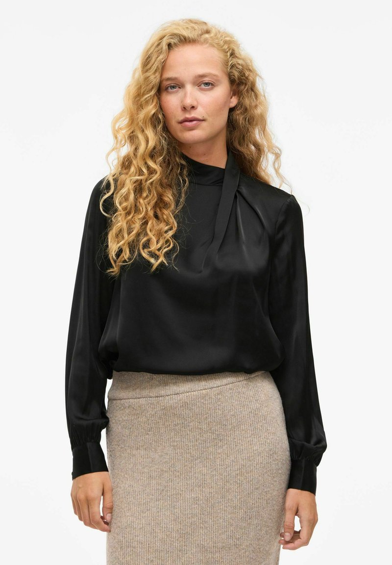 Blusa de satén negra con mangas largas y escote plegado, combinada con una falda beige texturizada. Tejido suave y corte relajado.